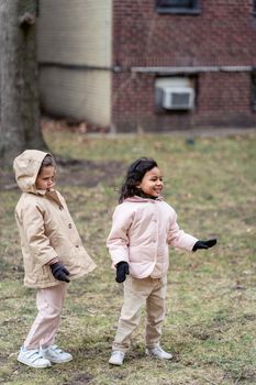 Cheerful Hispanic child with girlfriend in coat and gloves looking away on meadow in daytime
