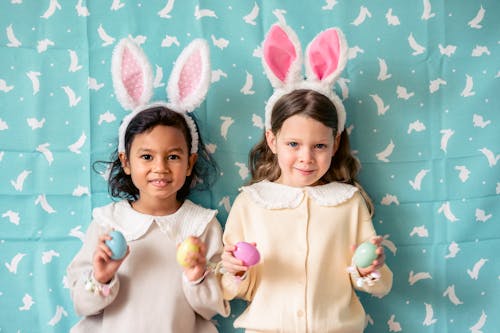 Free Smiling multiethnic children with bunny ears and plastic eggs looking at camera against fabric with rabbit pattern during festive event Stock Photo