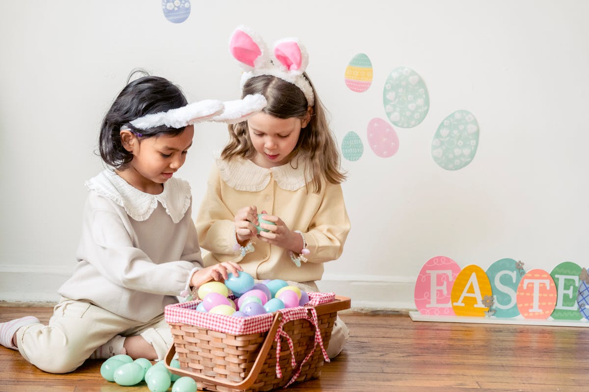 Best multiethnic friends sitting on parquet against basket with decorative eggs and Easter inscription at home