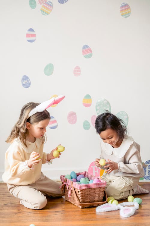 Charming girl with bunny ears sitting against smiling Hispanic child with decorative egg during festive occasion in house