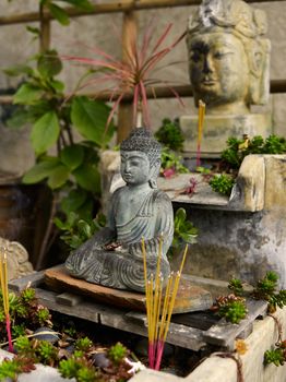 A serene Buddha statue with incense sticks is surrounded by vibrant plants in an outdoor shrine.