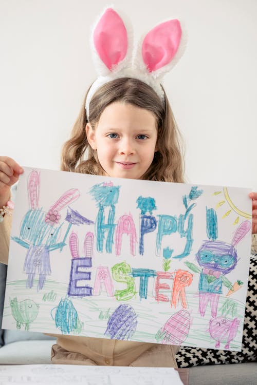 Child in headband with rabbit ears demonstrating paper drawing while looking at camera during festive occasion in house