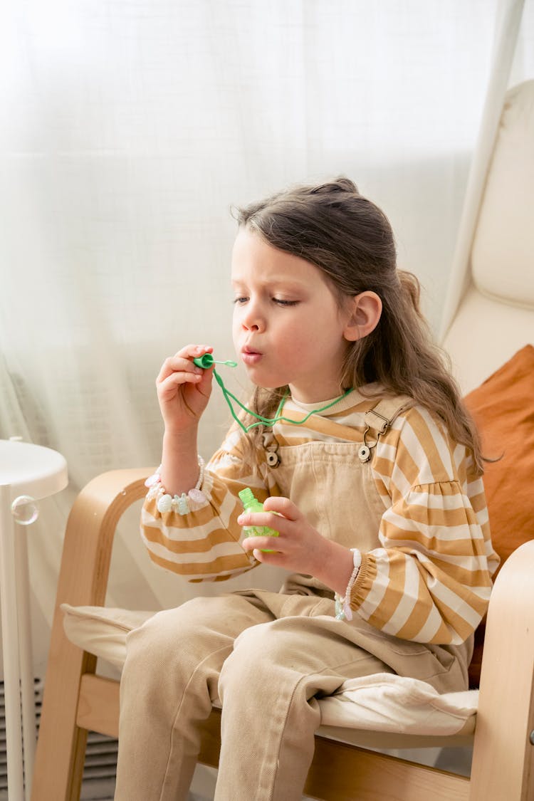 Girl Blowing Soap Bubbles In Rocking Chair At Home