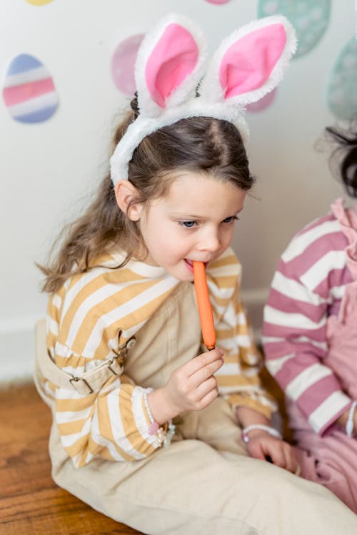 Child in headband with hare ears enjoying raw carrot while sitting against crop anonymous girlfriend and looking away on Easter Day