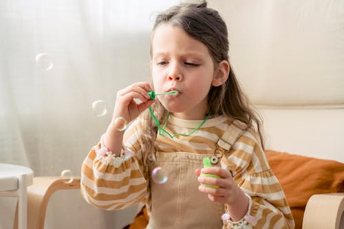 Attentive child in striped wear blowing soap bubbles while sitting in armchair in light house room