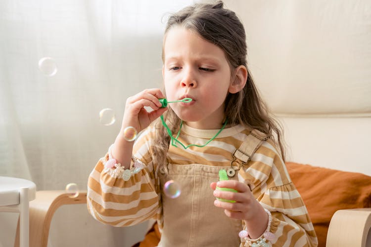 Girl Blowing On Soap Bubble Stick In Armchair