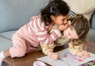 Attentive Hispanic child whispering in ear of girlfriend coloring paper drawing with crayon at table in house