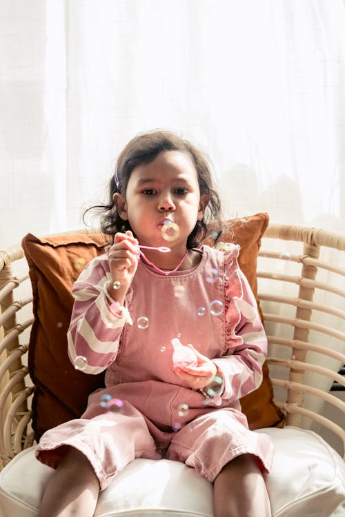 Hispanic girl blowing soap bubbles on chair in house