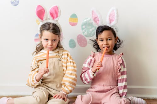 Multiethnic kids in headbands with hare ears eating fresh carrots while looking at camera on Easter Day at home