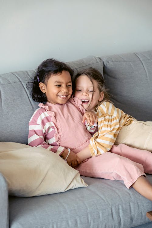 Cheerful child in striped wear embracing best ethnic friend while resting on sofa in living room