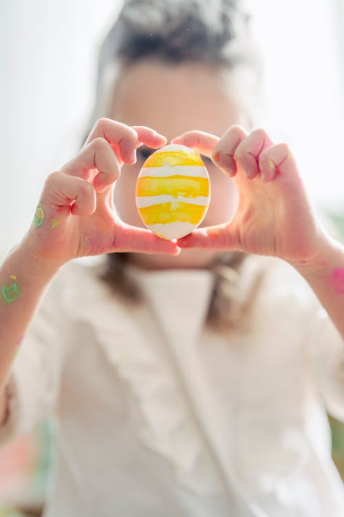 Anonymous child in white blouse with yellow painted Easter egg in hands in light room