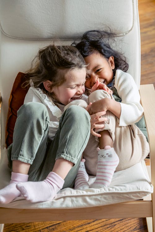 Adorable multiracial preschool girls laughing together while cuddling and sitting close on comfortable sofa
