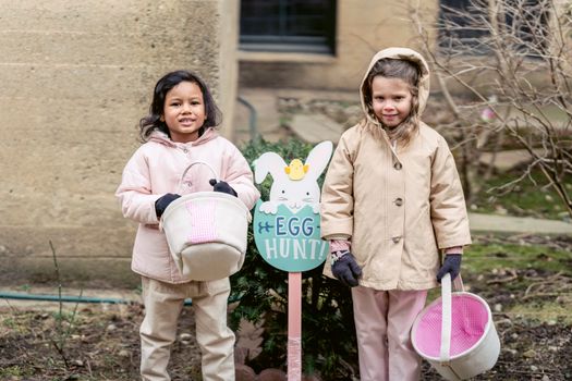 Multiracial girls in warm outfits looking at camera while standing with toy buckets near sign during egg hunting