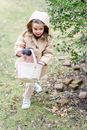 Little fair haired girl in warm bright coat and sneakers with toy bag walking on grass near tree outdoors