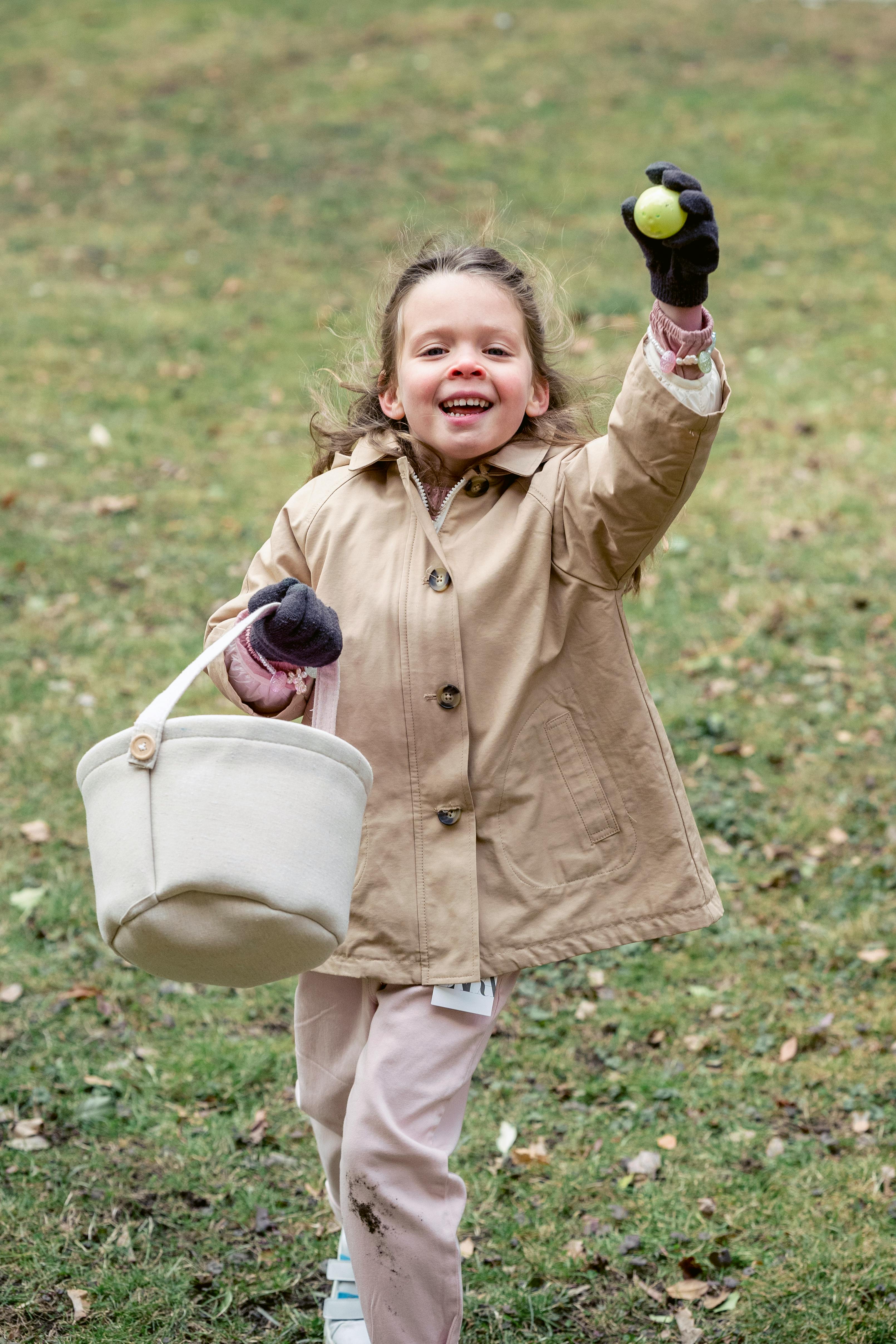 Adorable fair haired girl in warm coat with toy basket demonstrating green plastic egg while standing on grass