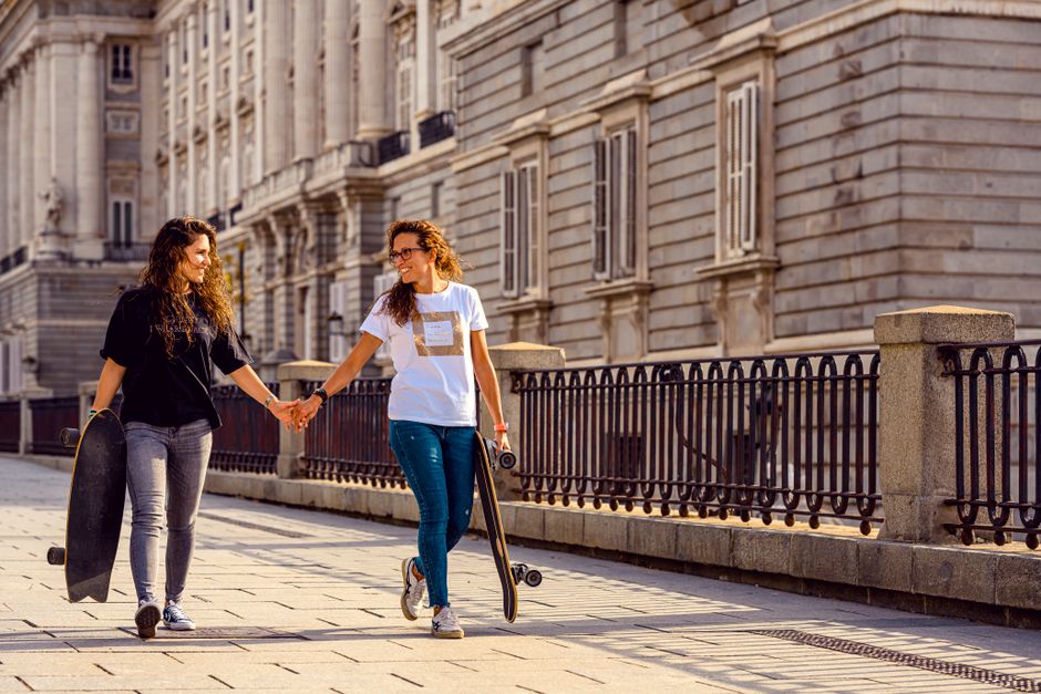 Dos mujeres de la mano con patinetes, mostrando amistad y diversión al aire libre.