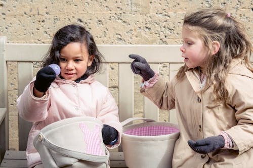 Free Adorable multiracial girls in outerwear sitting on bench with baskets while playing egg hunt game on street during Easter holiday Stock Photo