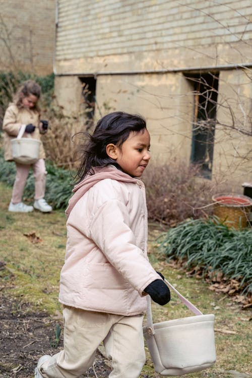 Free Hispanic preschool girl with basket playing game with friend while walking in backyard near building during Easter celebration on street Stock Photo