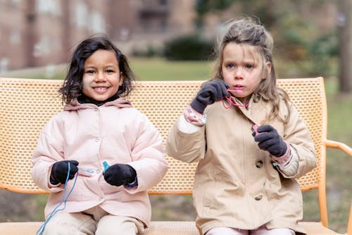 Free Positive diverse preschool girls in outerwear and gloves blowing soap bubbles while sitting on bench against residential buildings on blurred background Stock Photo