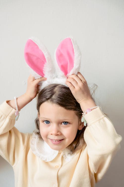 Adorable preschool girl in casual wear with bunny ears headband looking away while standing on white background in light room