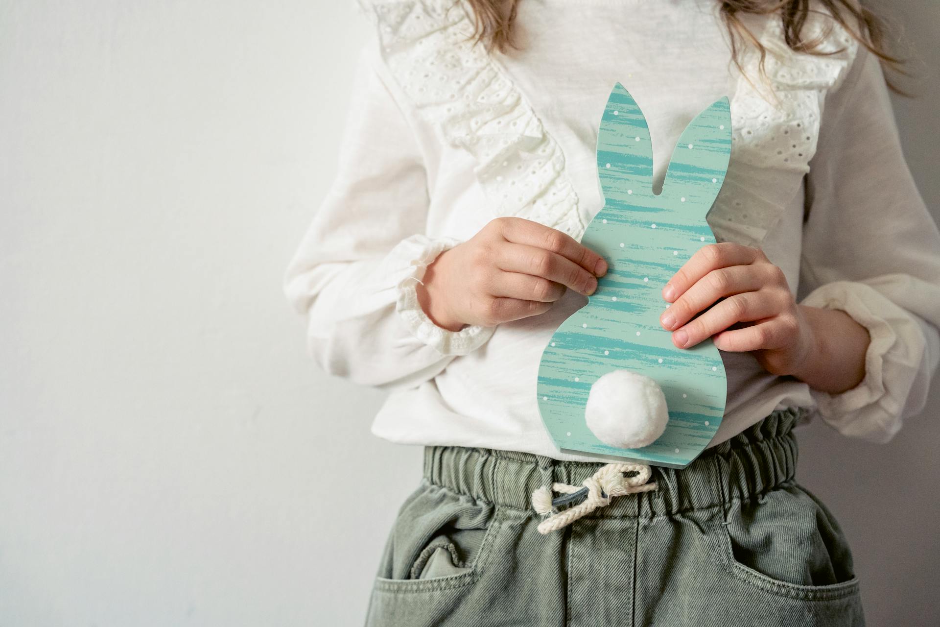 Crop unrecognizable girl with creative decoration in shape of bunny standing on white background during Easter holiday celebration in room