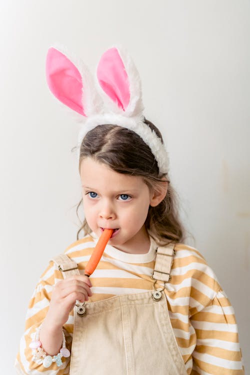 Cute little girl in stylish clothes and pink bunny ears eating sweet carrot during Easter holiday against white wall