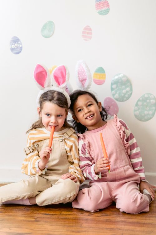 Free Adorable little multiracial girlfriends in stylish jumpsuits and bunny ears eating yummy sweet carrots while sitting together on floor near wall with decorative Easter eggs Stock Photo