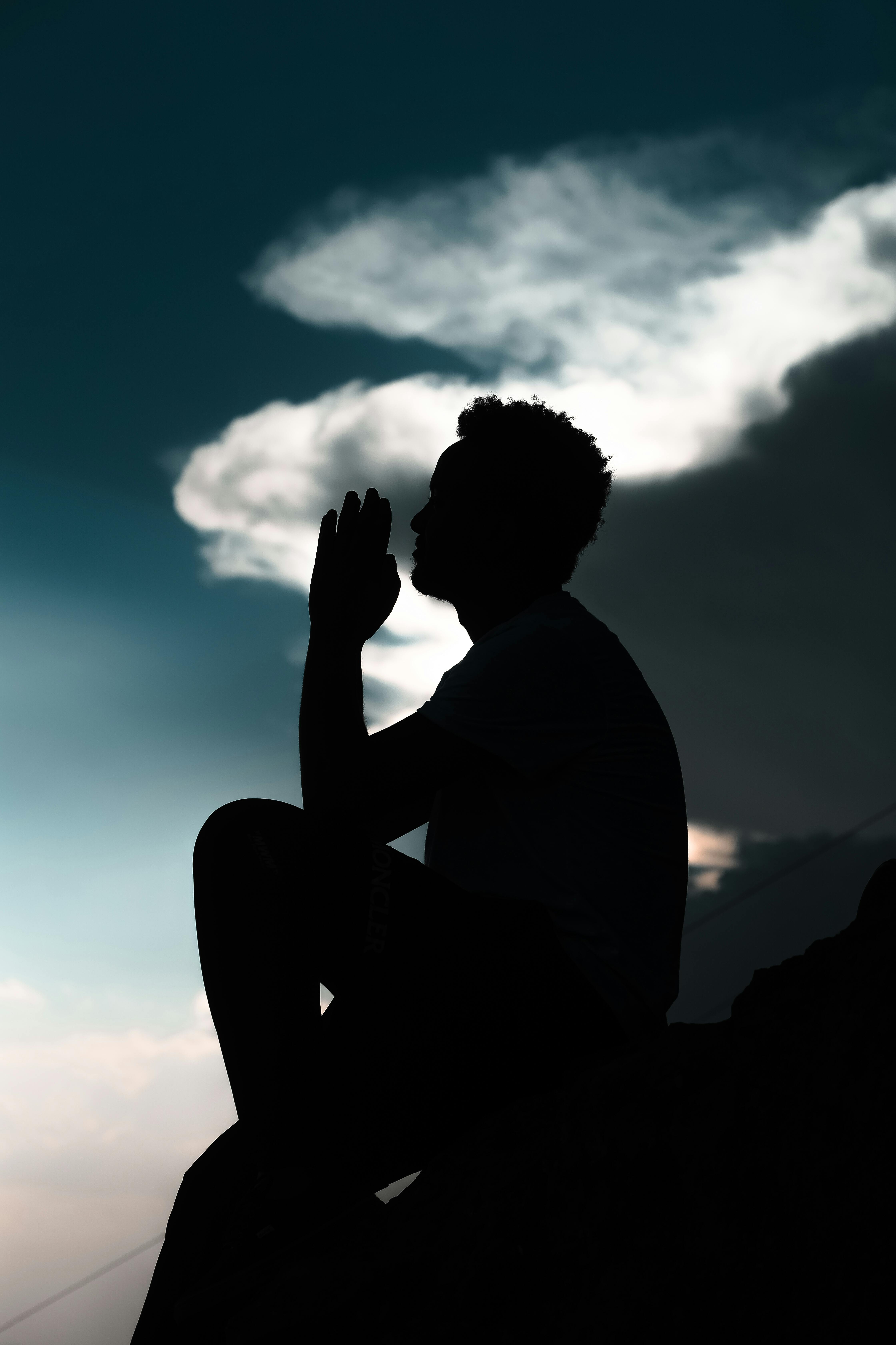 Silhouette of a man sitting and praying against a twilight sky with dramatic clouds.
