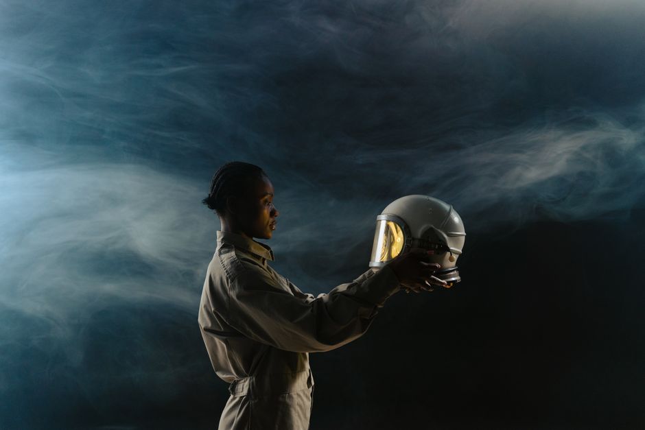 A woman stands in profile holding an astronaut helmet against a smoky backdrop.