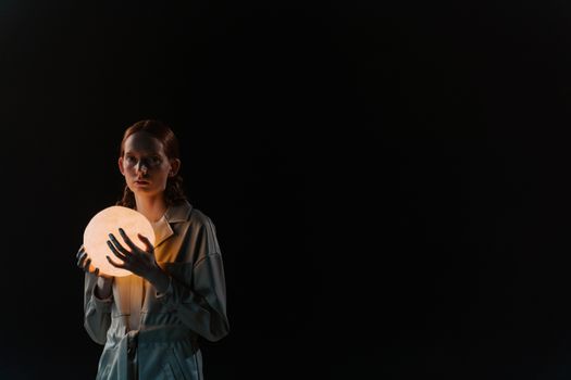 A woman in a trench coat poses with a glowing sphere against a dark background, creating a mysterious and artistic portrait.