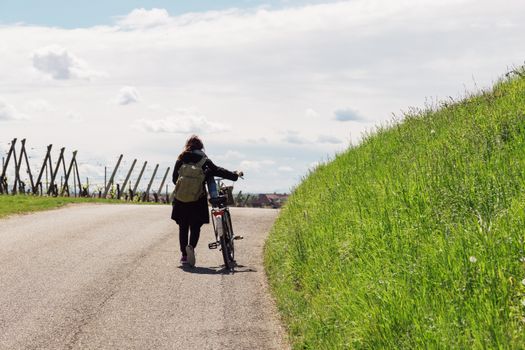 Woman walking with bicycle along a scenic country road with lush greenery and blue skies.