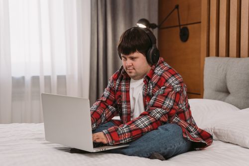Free A young man with headphones uses a laptop while sitting on a bed, embracing a relaxed lifestyle indoors. Stock Photo