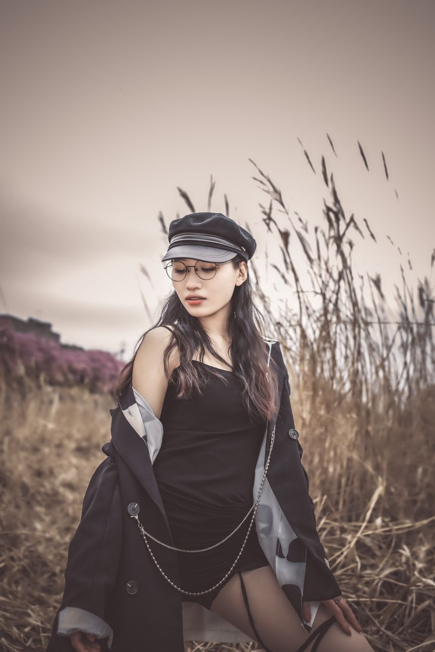 Fashionable young woman in a field, wearing a black hat and glasses, exuding a modern and edgy vibe.