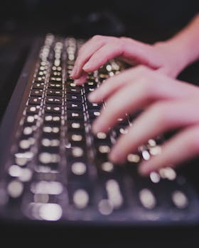Hands typing on an illuminated keyboard in a dimly lit room, showcasing technology and focus.