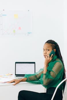 Young woman multitasking with phone and laptop at desk, focusing on work.