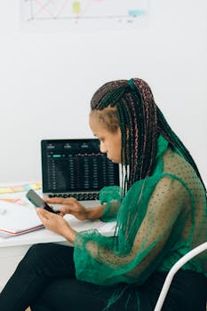 Businesswoman analyzing stock market data using smartphone and laptop indoors.