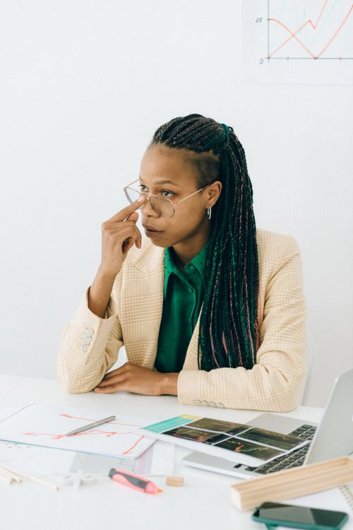 Free African American woman with braided hair in glasses focusing on work tasks at a desk with charts and a laptop. Stock Photo