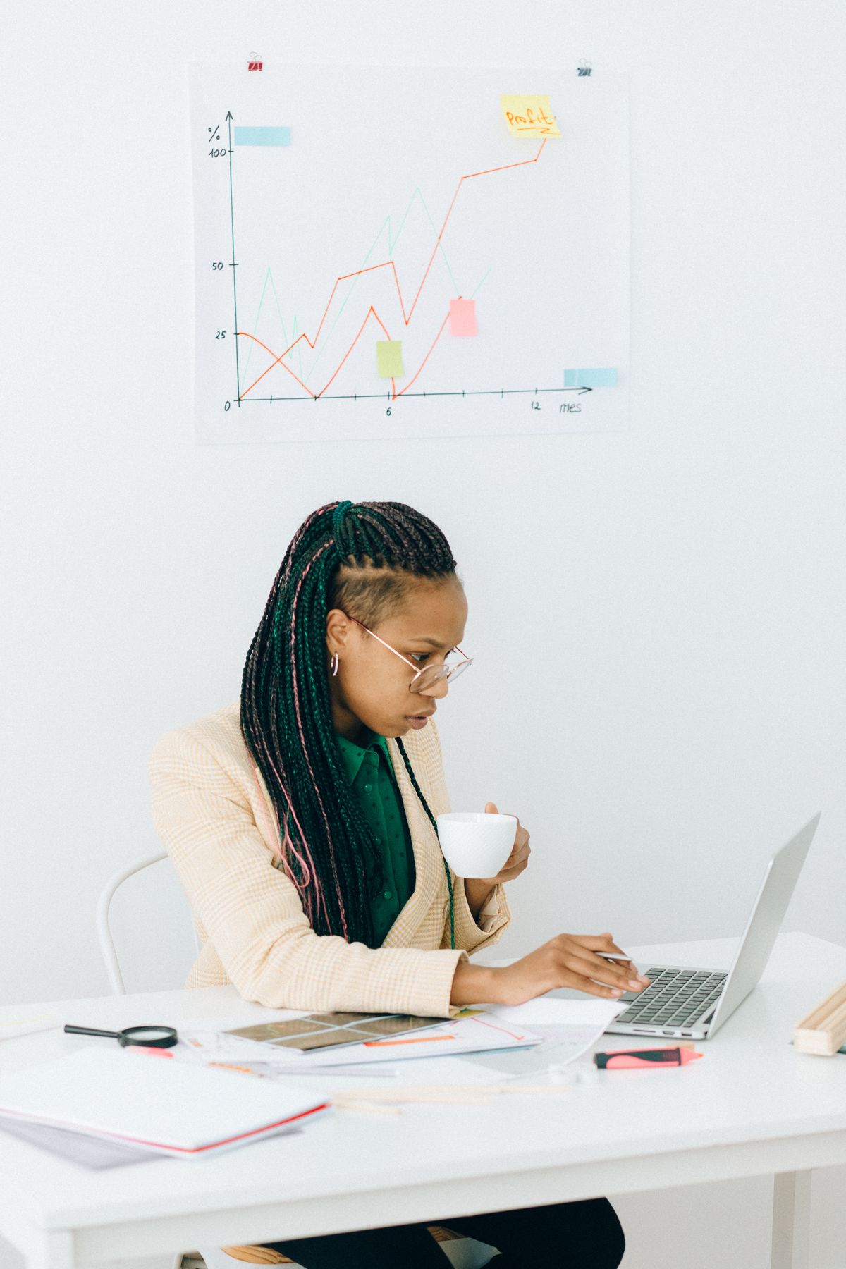 A Black woman in a beige blazer sits at a white desk with a coffee cup, working at a laptop; printed charts, papers, a magnifying glass, and a wall chart with sticky notes are visible across her workspace.