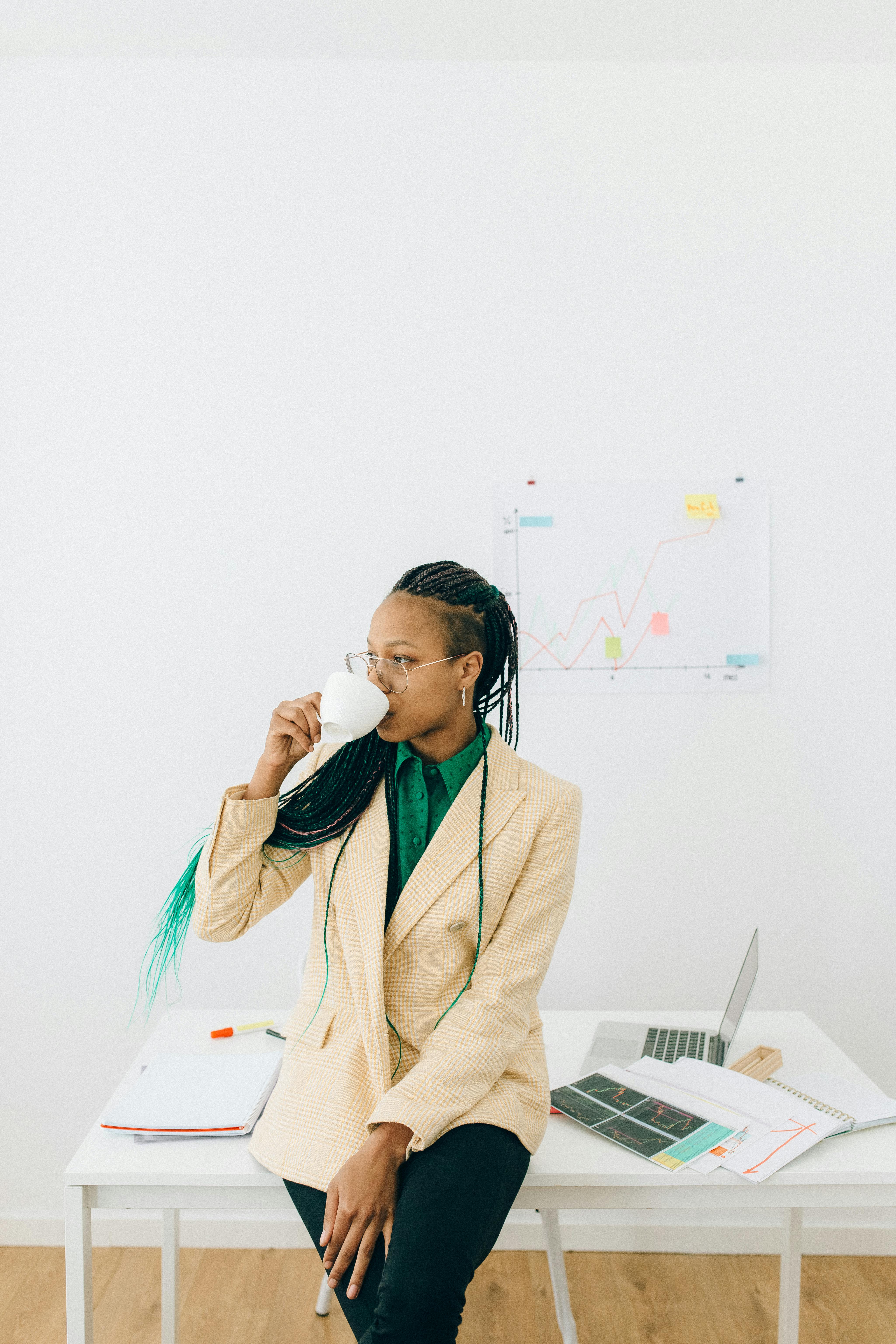 woman in beige coat drinking from a white cup in front of her work desk