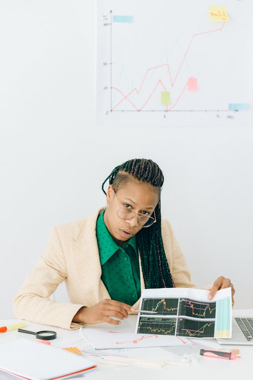 Free Woman in Beige Coat Pointing at the Graph in the Paper while Looking at the Camera Stock Photo