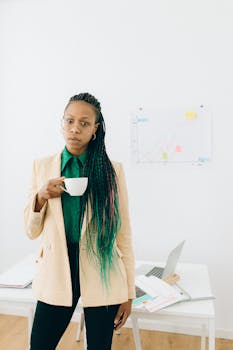 Confident professional woman with braids holding coffee in modern office.