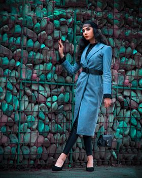 Elegant woman in a blue coat posing against a stone wall with a handbag, showcasing fashion and style.