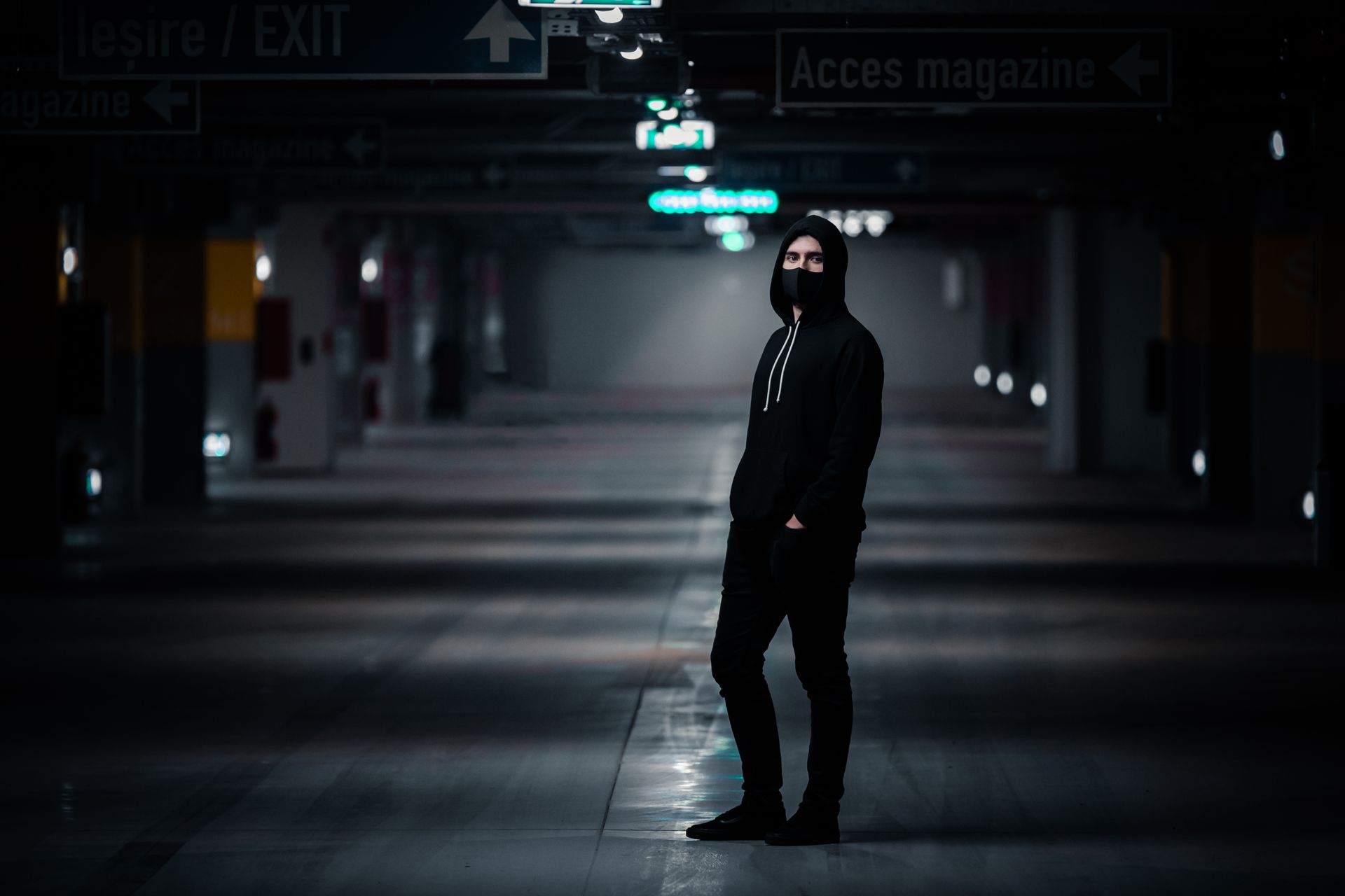 A man in a black hoodie stands in an atmospheric underground parking garage, creating a mysterious urban portrait.