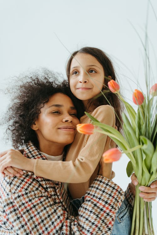 Woman in Checked Coat Hugging a Child while Holding a Bunch of Flowers