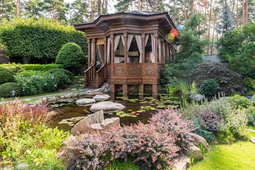 Free Aged wooden summer house near pond with rocks near green plants and flowers in garden in sunny day Stock Photo