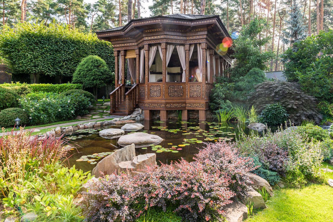 Aged wooden summer house near pond with rocks near green plants and flowers in garden in sunny day