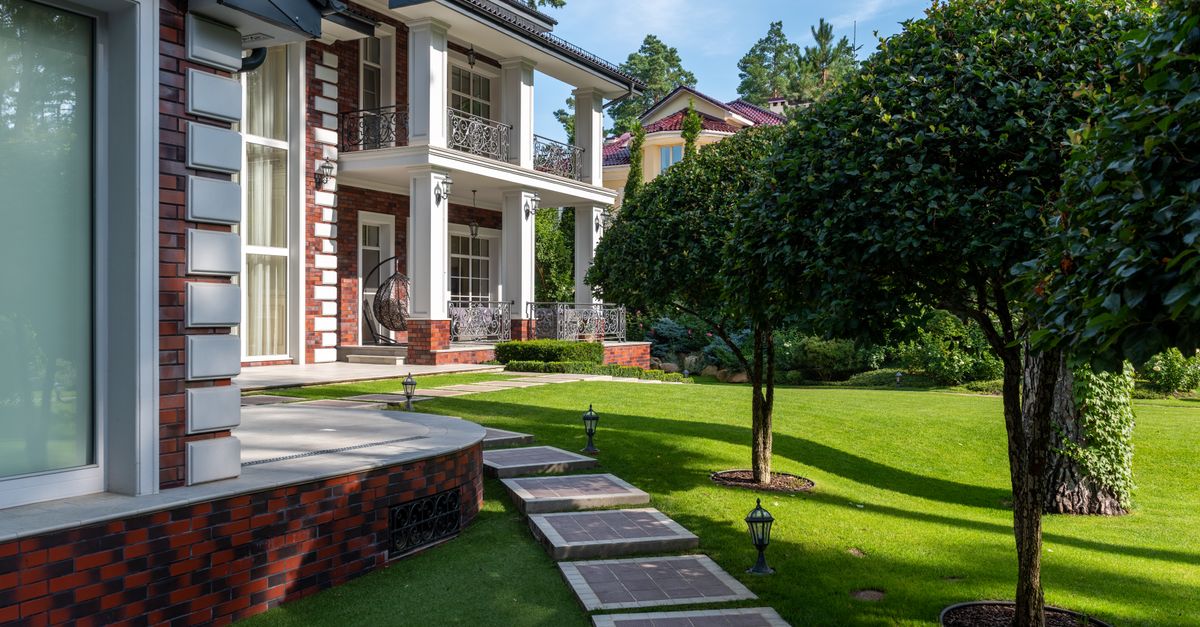 Modern house near backyard with path near green grass and plants with trees under blue sky in summer sunny day