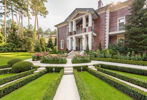 Luxurious brick mansion surrounded by lush gardens and trimmed hedges on a sunny day.