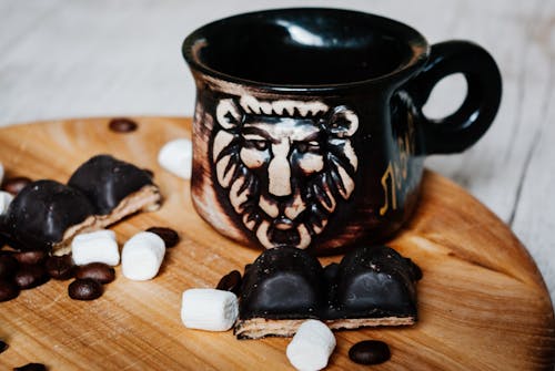 Brown shiny fragile mug with lion placed on wooden table with soft sweet white marshmallows and chocolate waffles