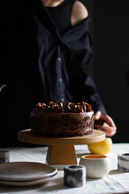 Crop anonymous female standing at table with sweet chocolate cake and plated near candle holders